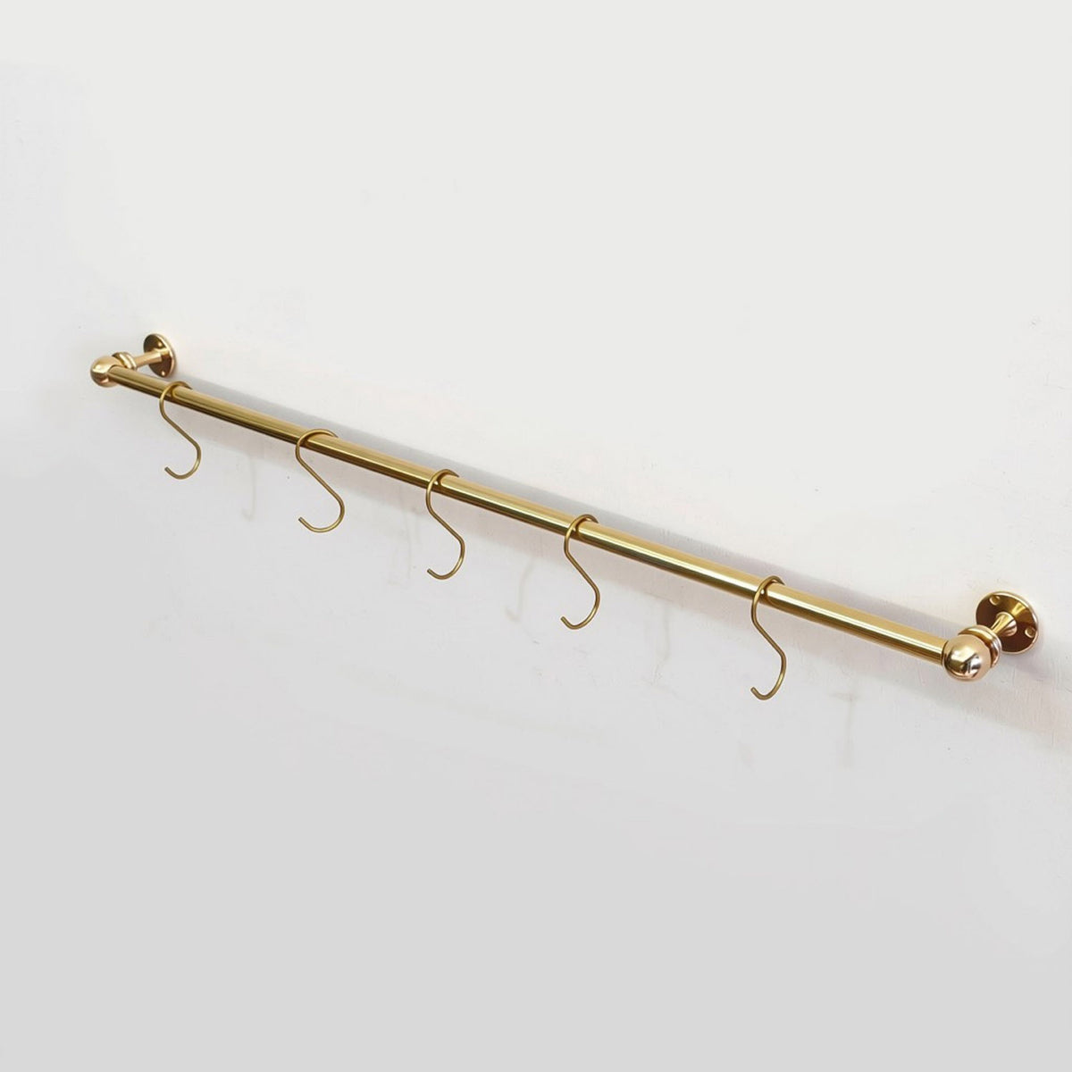 Front-angle view of a compact solid unlacquered brass kitchen hanging rod with five S-hooks mounted on a white wall.