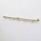 Angled full view of a long wall-mounted unlacquered solid brass kitchen rail with six S-hooks against a plain white background.