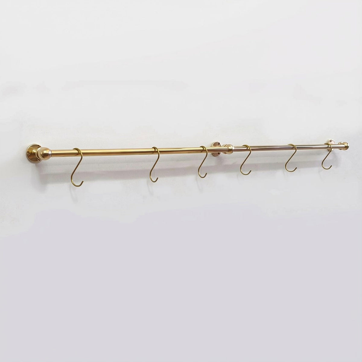 Full view of a long solid unlacquered brass kitchen hanging rail mounted on a plain white wall with six brass S-hooks.