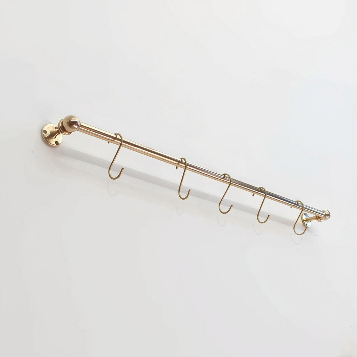 Perspective view of a short solid unlacquered brass kitchen hanging rail mounted on a plain white surface with five matching S-hooks.