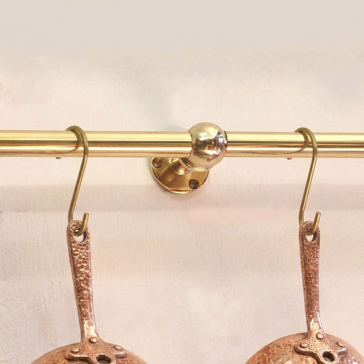 Center support bracket detail of a solid unlacquered brass kitchen rail with S-hooks holding copper cookware on a white wall.